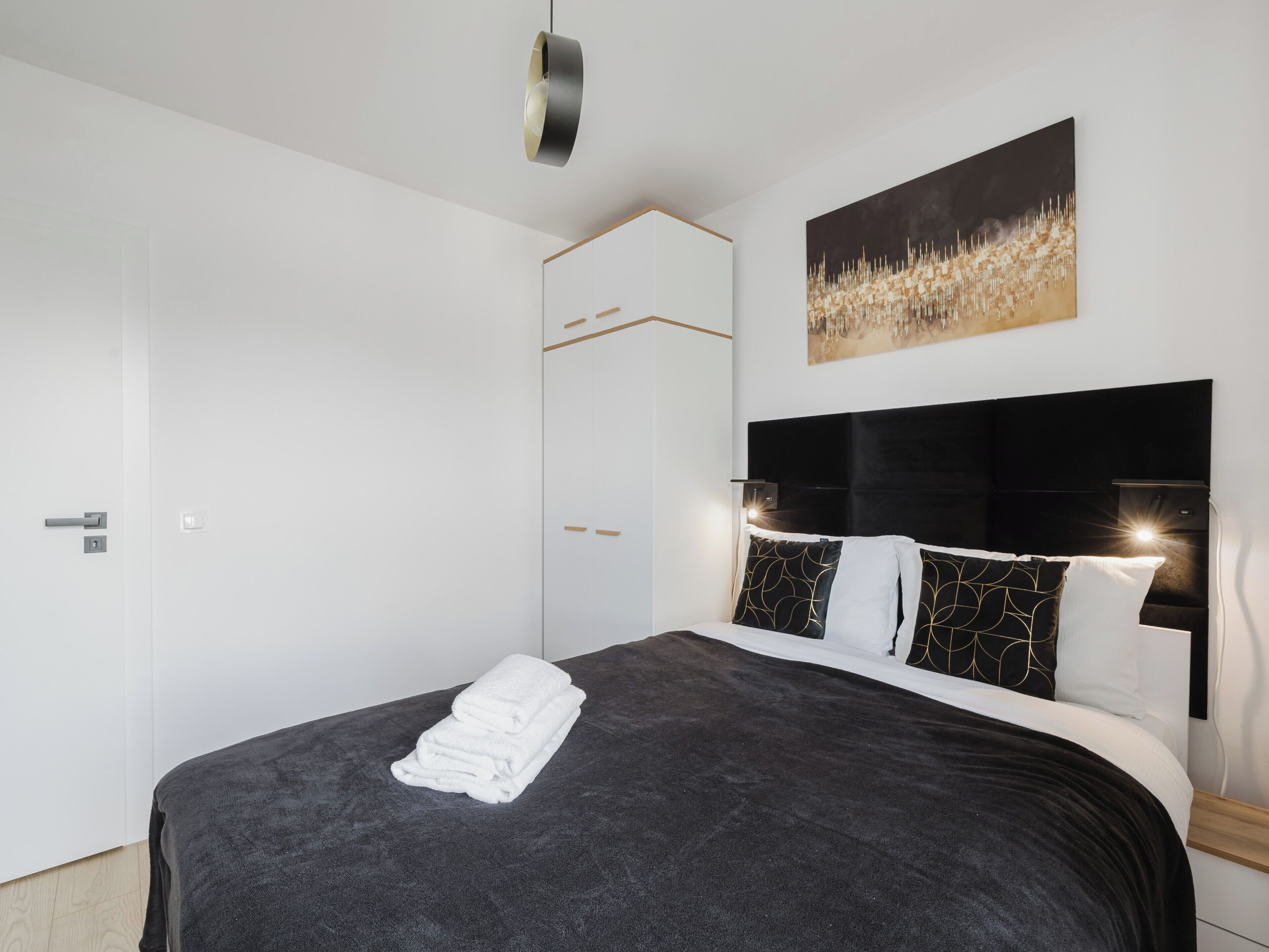 A modern bedroom with a large bed, a headboard with integrated lighting, a painting on the wall, and a wardrobe with a mirror.