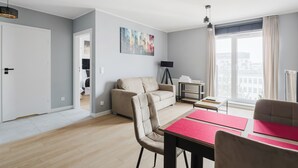 A modern living room with a sofa, dining table, and chairs. There is a painting on the wall, a floor lamp, and a window with curtains. - Fiqus Marcelin Deluxe Apartment (Poznan)