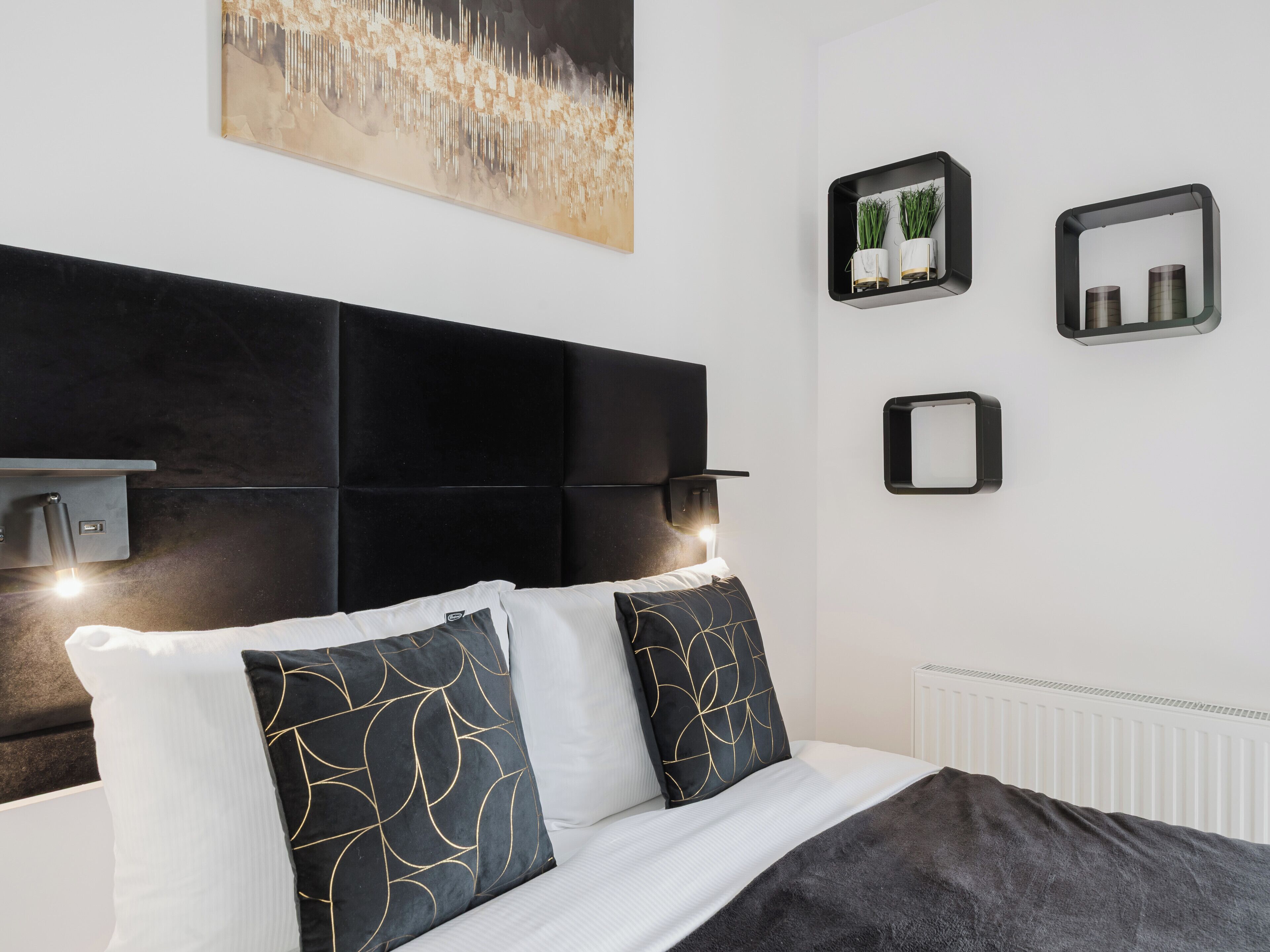 Une chambre à coucher moderne avec une tête de lit noire, du linge de lit blanc et des coussins décoratifs. On y trouve des étagères murales ornées de plantes et un tableau au-dessus du lit.