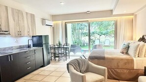 A modern living room with a sofa, armchairs, and a dining area with a table and chairs. There is a kitchen area with cabinets and a refrigerator. The room has a large sliding glass door leading to an outdoor area with a view of greenery. - Coronado Luxury Club & Suites - swim up with golf for 2 and resort amenities! (Panamá Oeste Province)