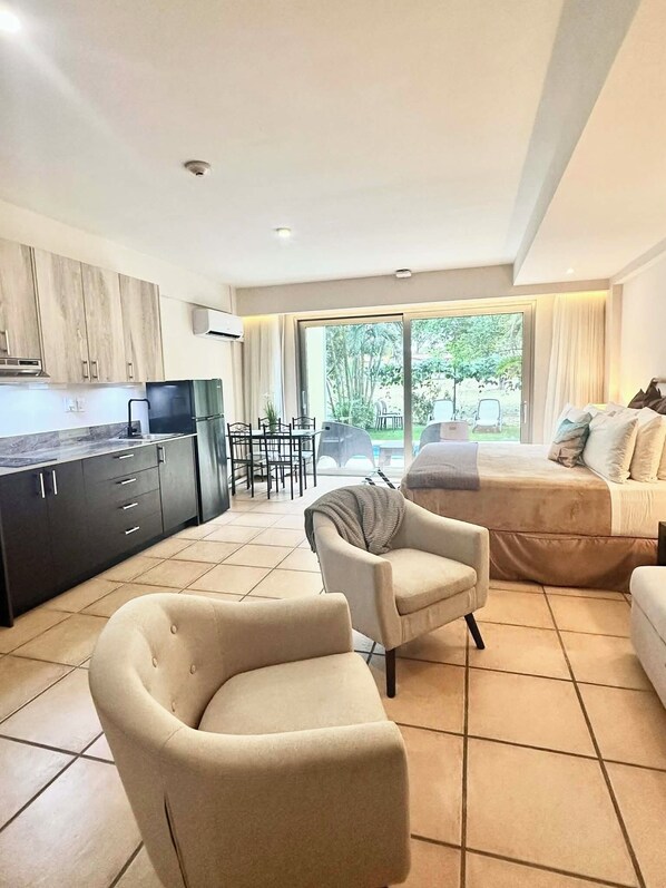 Een moderne woonkamer met een bank, fauteuils en een eethoek met een tafel en stoelen. Er is een keuken met kastjes en een koelkast. De kamer heeft een grote schuifpui die uitkomt op een buitenruimte met uitzicht op groen.
