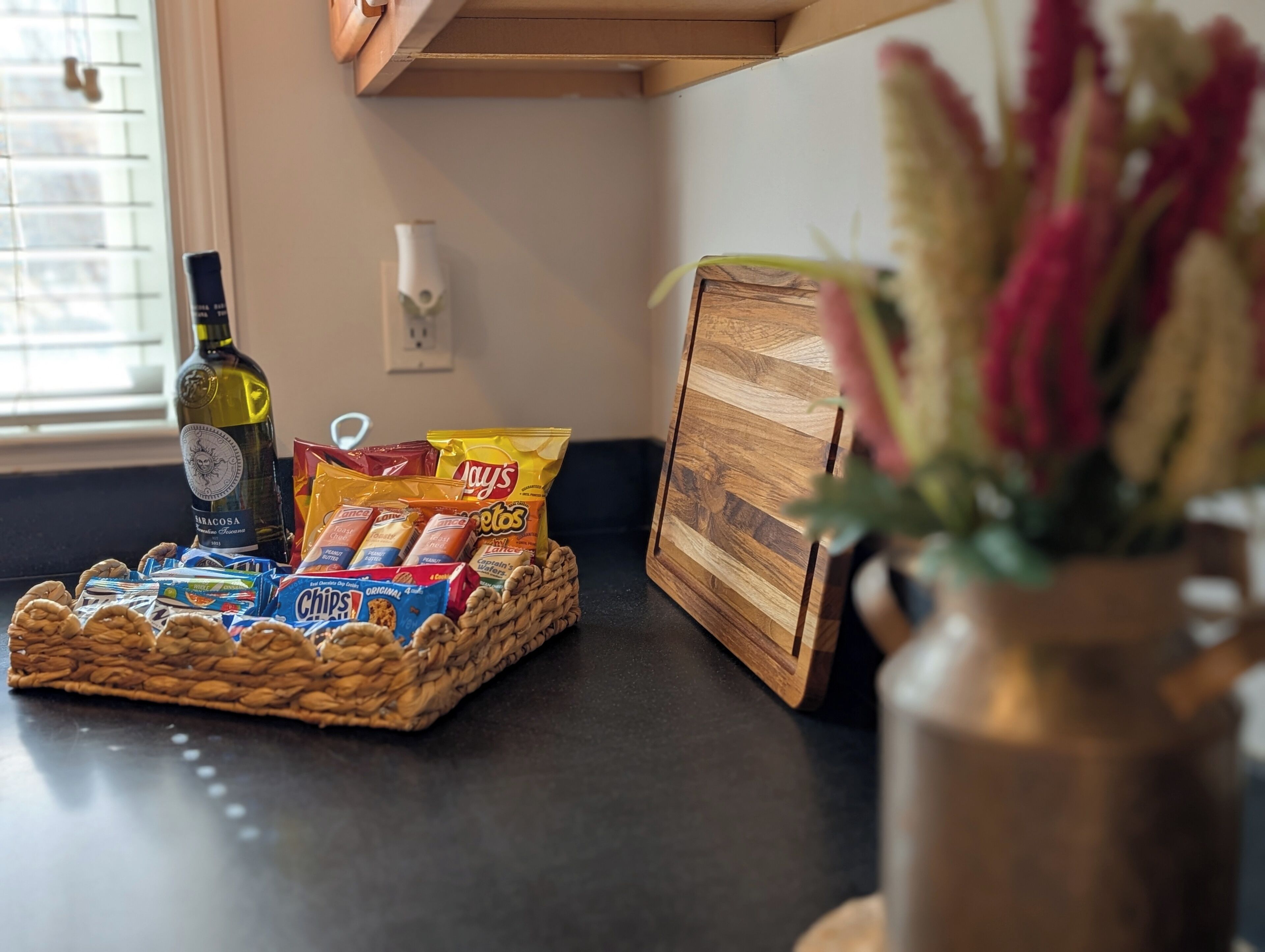 A kitchen counter with a basket of snacks, a bottle of olive oil, and a wooden cutting board.