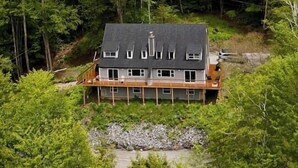 Ein Haus mit dunklem Dach, vielen Fenstern und einer Holzterrasse, umgeben von dichtem Grünwald.