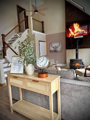 Ein Wohnzimmer mit einem Holztischchen, einer Zimmerpflanze und zwei Uhren. Im Hintergrund sind eine Treppe und ein Kamin mit einem Fernseher zu sehen.