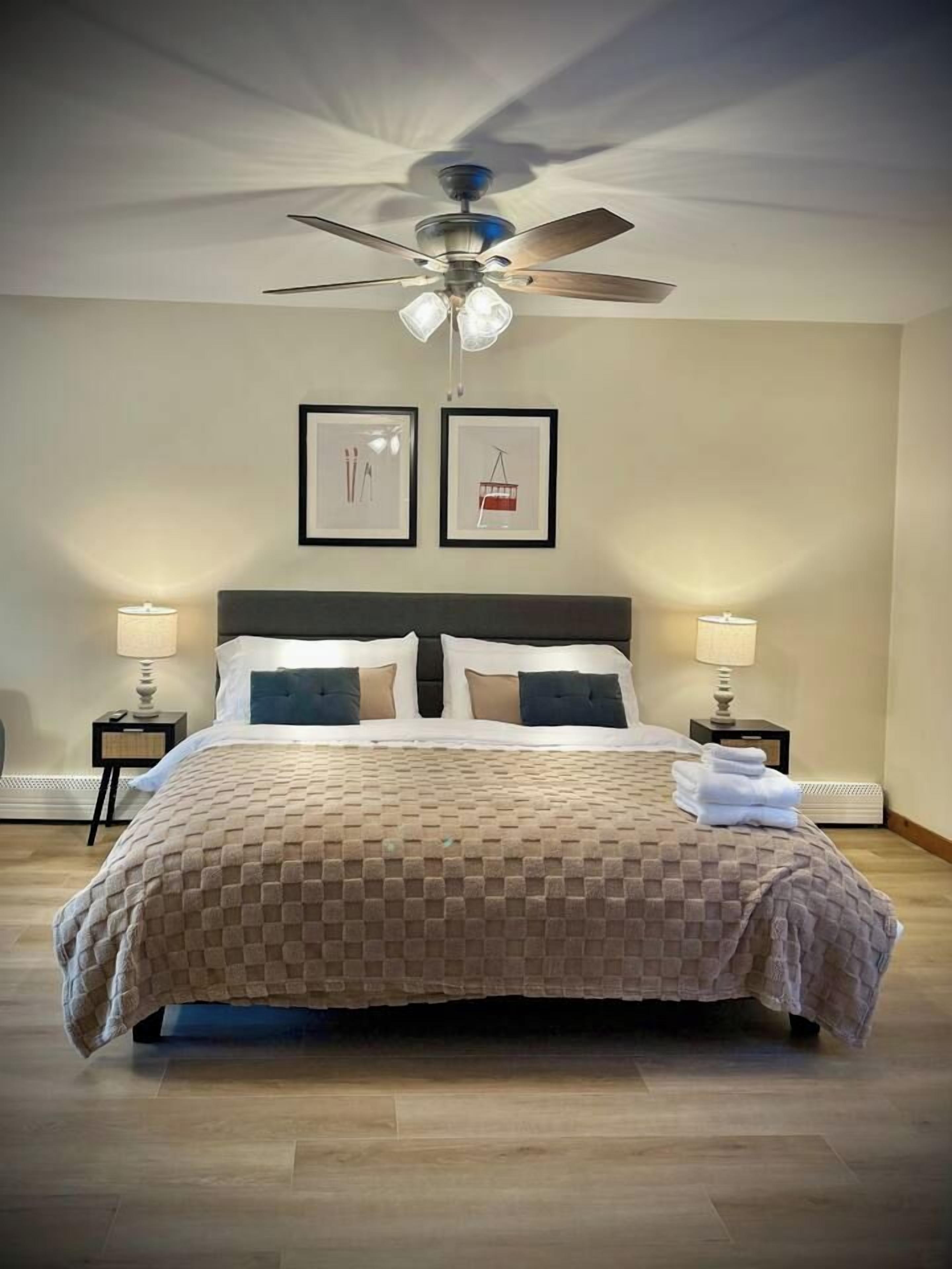 A neatly made bed with a checkered bedspread, two bedside lamps, a ceiling fan, and two framed pictures on the wall.