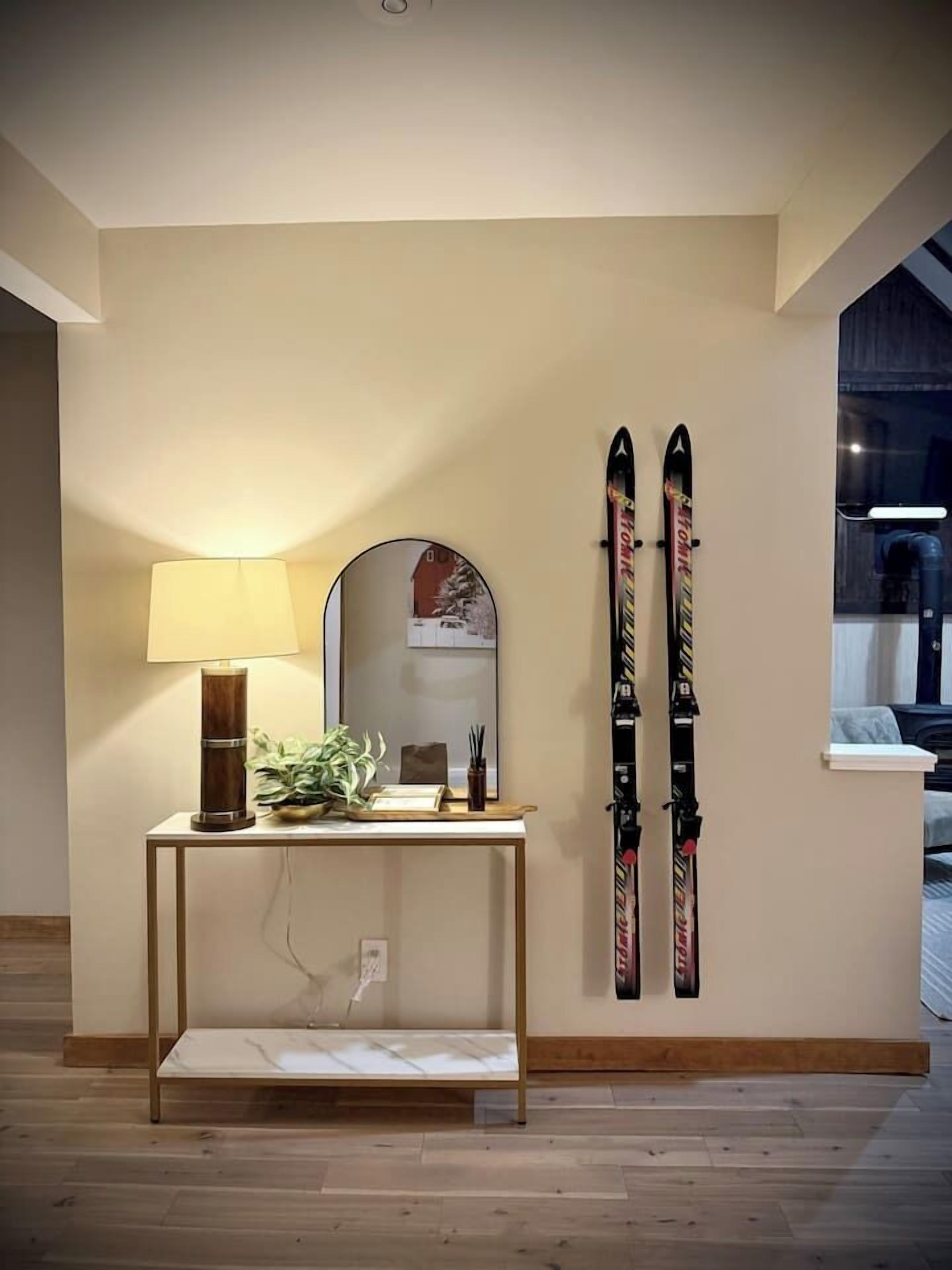 A room with a wooden floor, a small console table with a lamp, a mirror, and skis leaning against the wall.