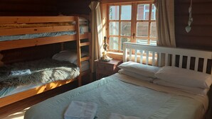 A wooden cabin room with bunk beds and a single bed, a window with curtains, and a small table with a lamp. - Small rustic log cabin, centrally located on the Isle of Skye. (Broadford)