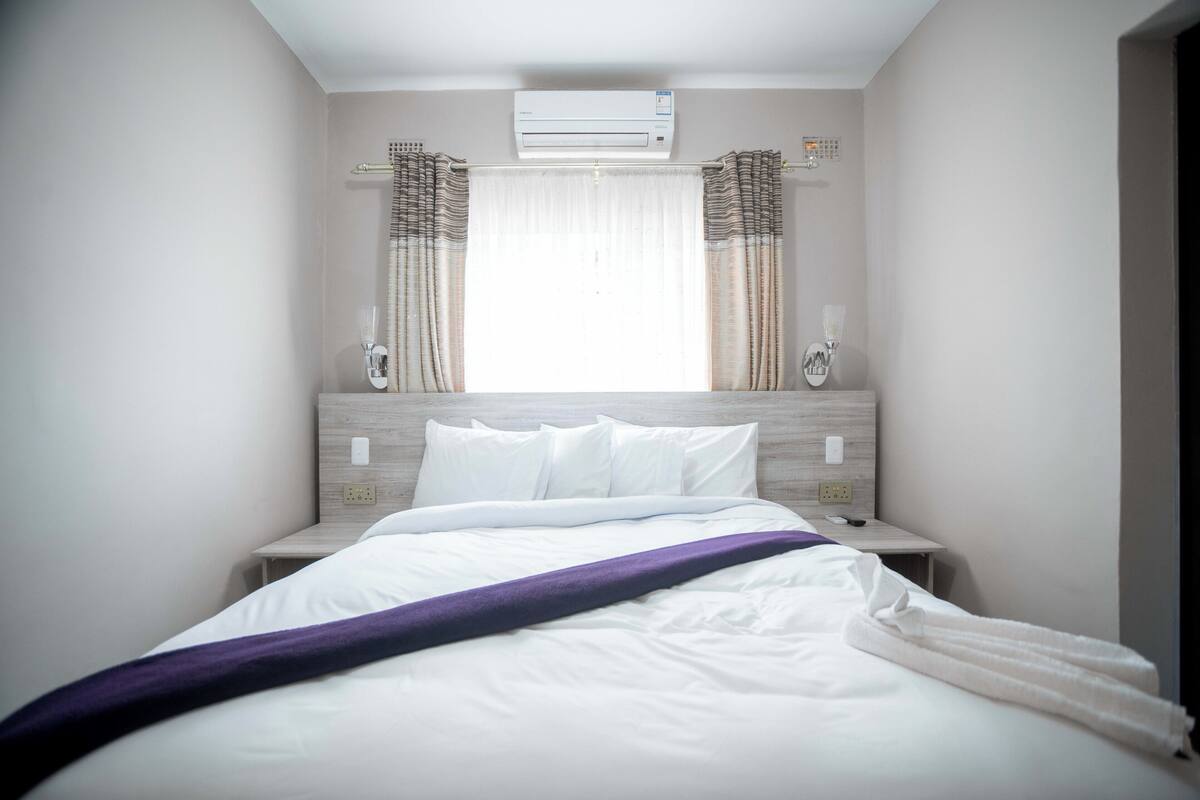 A hotel room with a bed, a nightstand, a window with curtains, and an air conditioning unit.