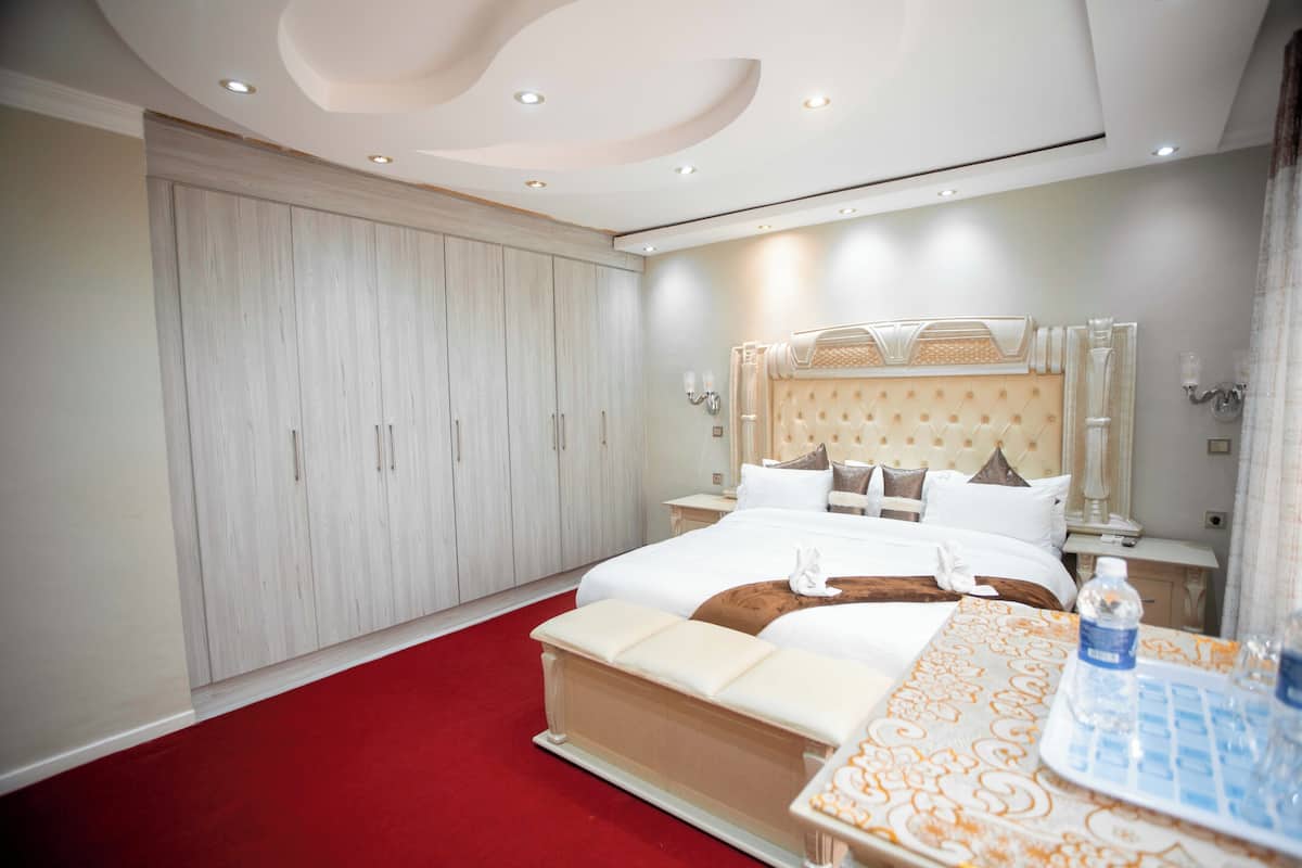 A hotel room with a large bed, a red carpet, a wooden wardrobe, and a bedside table with a bottle and glasses.