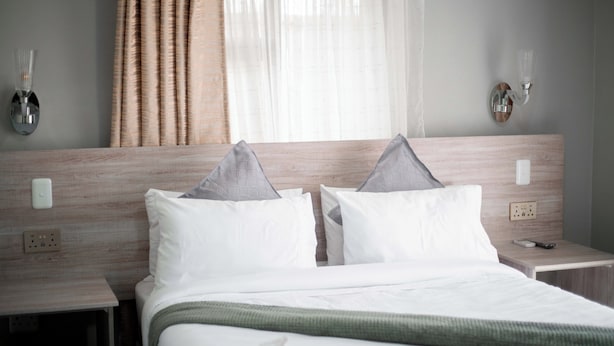 Habitación de hotel con cabecera de madera, dos almohadas blancas, una manta verde, una mesita de noche con lámpara y una ventana con cortinas traslúcidas.