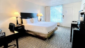 Quarto de hotel com uma cama, duas luminárias de cabeceira, uma escrivaninha com uma placa indicando "NEW YORK", uma cafeteira e uma porta.