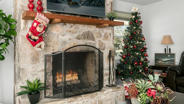 A stone fireplace with a TV above it, a Christmas tree, and a mantel with a stocking and candles. - Karen's Kottage | 3/2, Hot Tub, Fireplace, Fire-pit & Grill, Man Cave w/ Games + Bar (Fredericksburg)