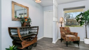 A room with a wooden cart holding wine glasses, a floral armchair, a mirror, and a window with a view of a house. - Karen's Kottage | 3/2, Hot Tub, Fireplace, Fire-pit & Grill, Man Cave w/ Games + Bar (Fredericksburg)