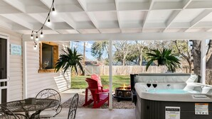 A covered patio area with a hot tub, red chairs, a metal table with chairs, and a sign indicating the lounge bar is now open. - Karen's Kottage | 3/2, Hot Tub, Fireplace, Fire-pit & Grill, Man Cave w/ Games + Bar (Fredericksburg)