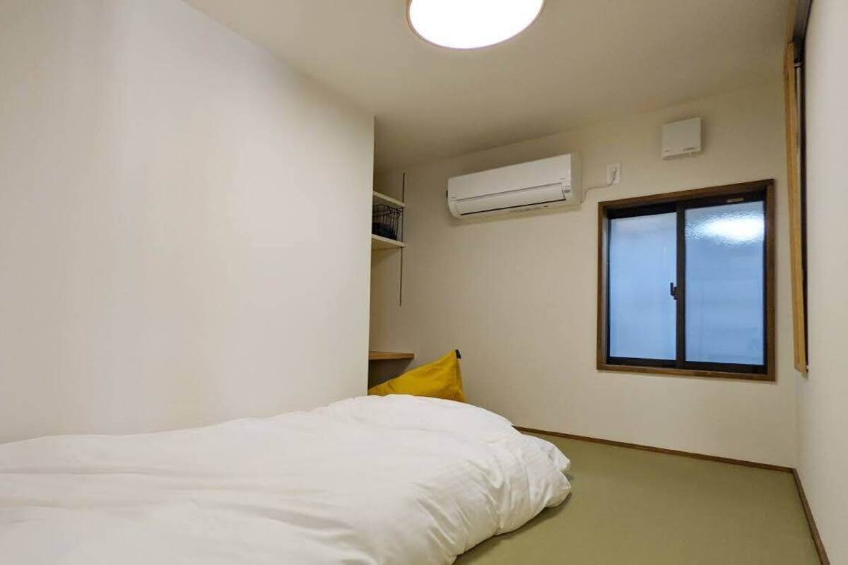 A small, sparsely furnished room with a bed, a window, and an air conditioning unit on the wall.