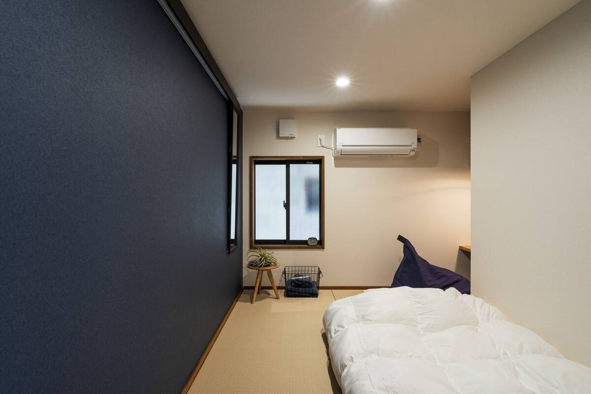 A bedroom with a bed, a window, an air conditioner, and a small plant on a stool.