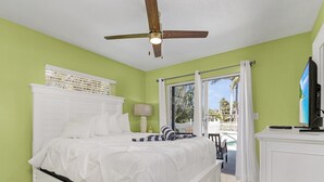 Une chambre à coucher avec un lit blanc, un ventilateur de plafond, une fenêtre avec des rideaux et une vue sur l’extérieur.