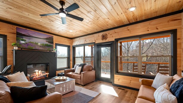 A cozy cabin interior with a fireplace, leather sofas, wooden floors, and large windows offering a view of the outdoors. - NEW, INDOOR POOL, GAME ROOM, BUNK ROOM, HOT TUB (Sevierville)