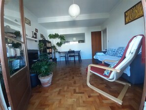 Ein Wohnzimmer mit Holzdielenboden, einem roten und weißen Schaukelstuhl, einer Couch mit gemusterten Kissen, einem Esstisch mit Stühlen und einer großen Topfpflanze.