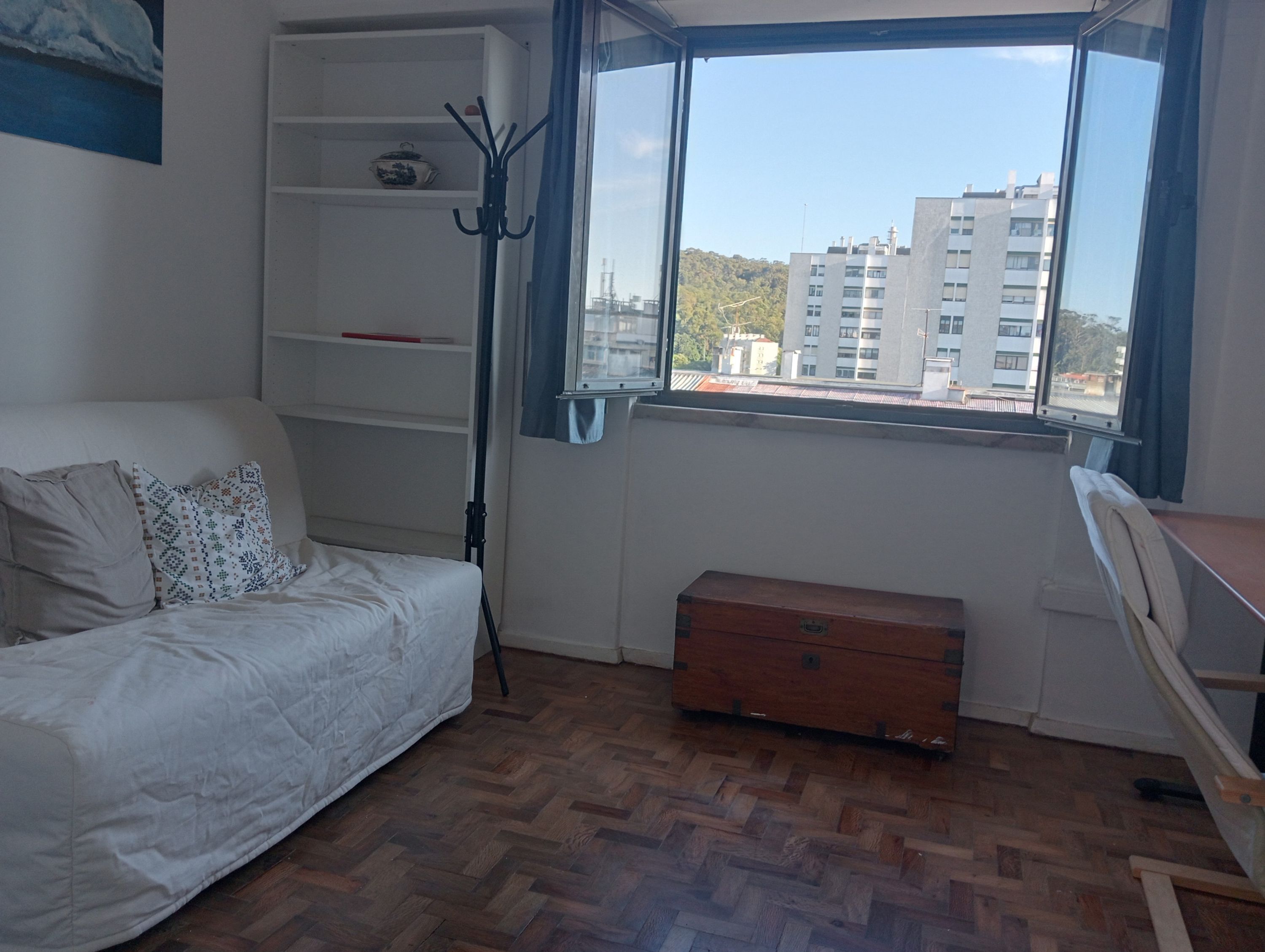 A room with a bed, a wooden chest, a desk, and a window with a view of buildings.