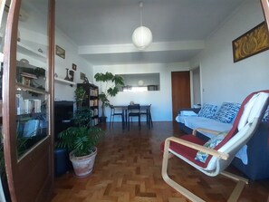 Ein Wohnzimmer mit Holzboden, einer Couch, einem Esstisch mit Stühlen, einem Bücherregal und einem Blumentopf.