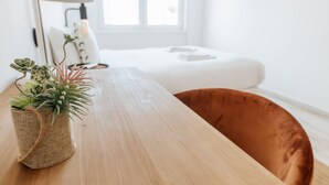 A wooden table with a woven basket containing green plants, a white lamp, and a white bed with a brown headboard in a bright room with a window.