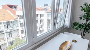 A room with a white table, a plant, and a view of buildings through large windows.