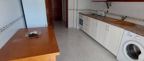 Uma cozinha compacta com armários brancos, forno embutido, máquina de lavar e mesa de madeira.