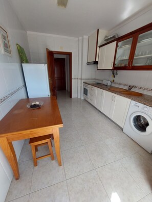 A compact kitchen with white cabinets, a built-in oven, a washing machine, and a wooden table.