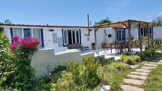 A white building with blue trim, a patio with a wooden table and chairs, and a garden with various plants and flowers. - Beach Wave Melides Villa (Grandola)