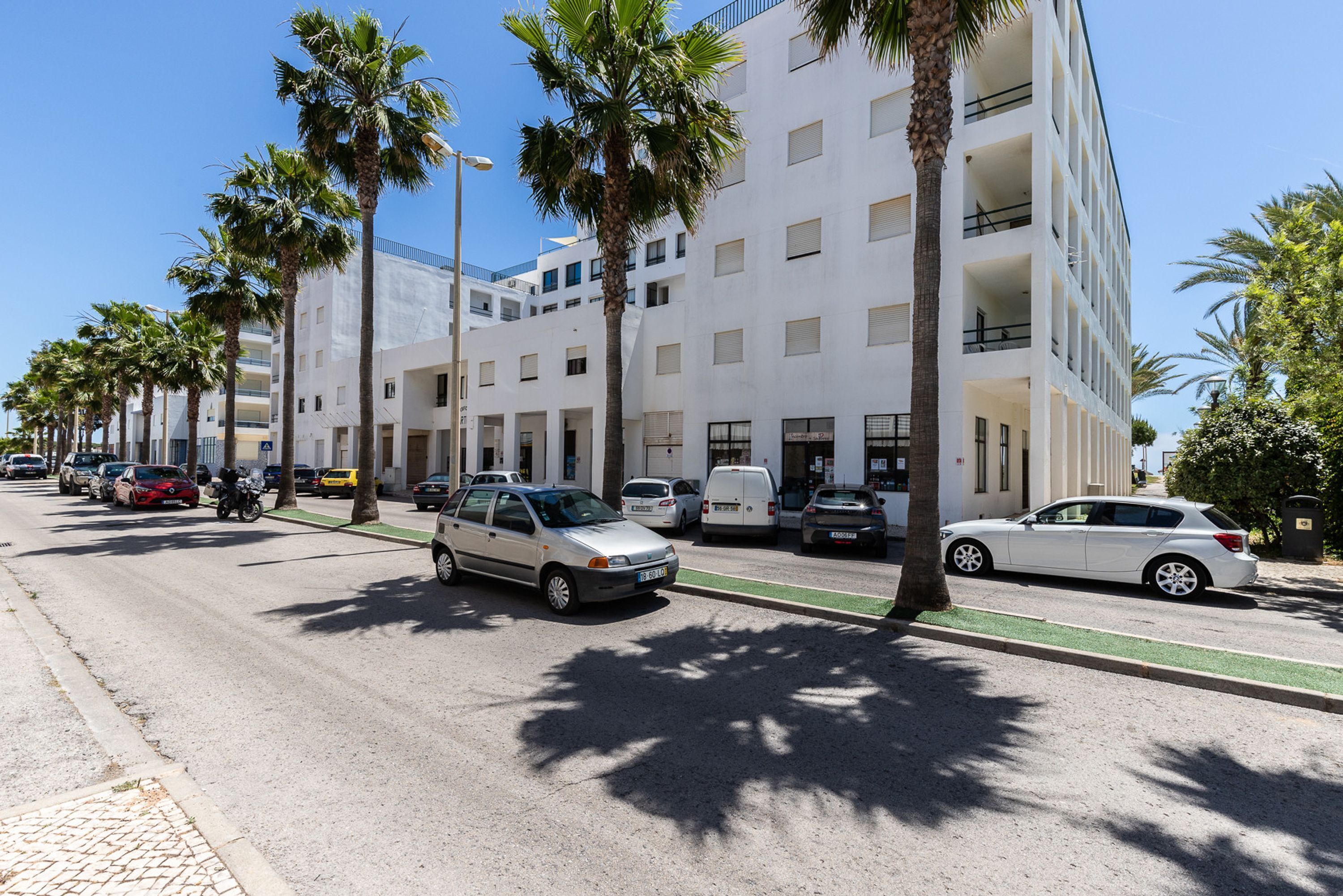 Un edificio moderno con diversi piani, balconi e una strada con auto parcheggiate e palme.