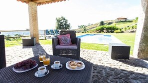 Un espacio exterior con mobiliario de mimbre, una mesa con una fuente de uvas, tazas de café y un vaso de zumo de naranja.