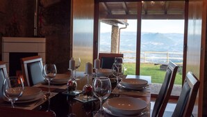 A dining table set for six with wine glasses, plates, and cutlery. The room has a stone wall and a large window with a view of mountains and a body of water. - Luxury Villa Private Pool Belle Poque (Celorico de Basto)