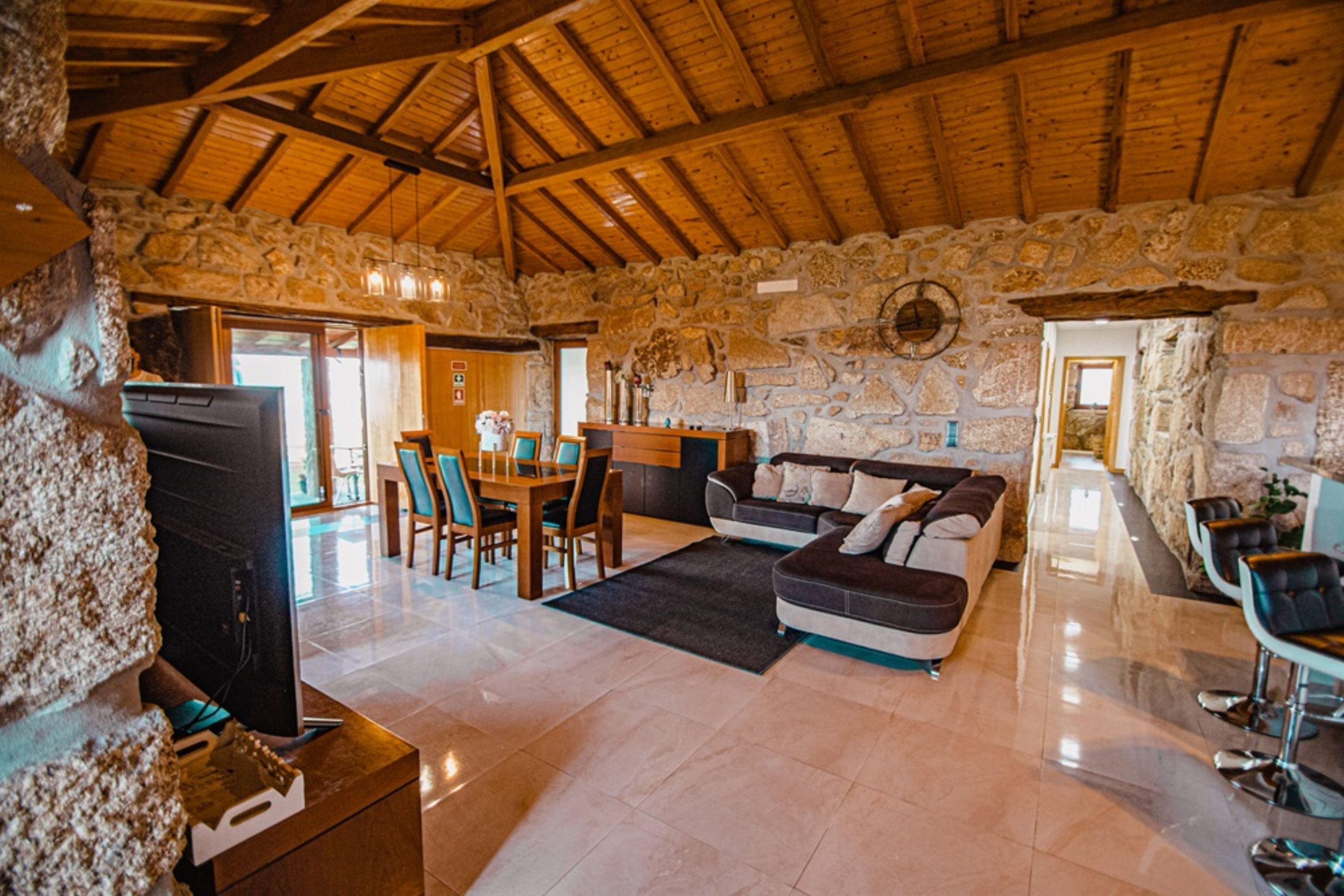 A spacious living area with a stone wall, wooden ceiling, a sofa, a TV, and a dining area with chairs and a table.