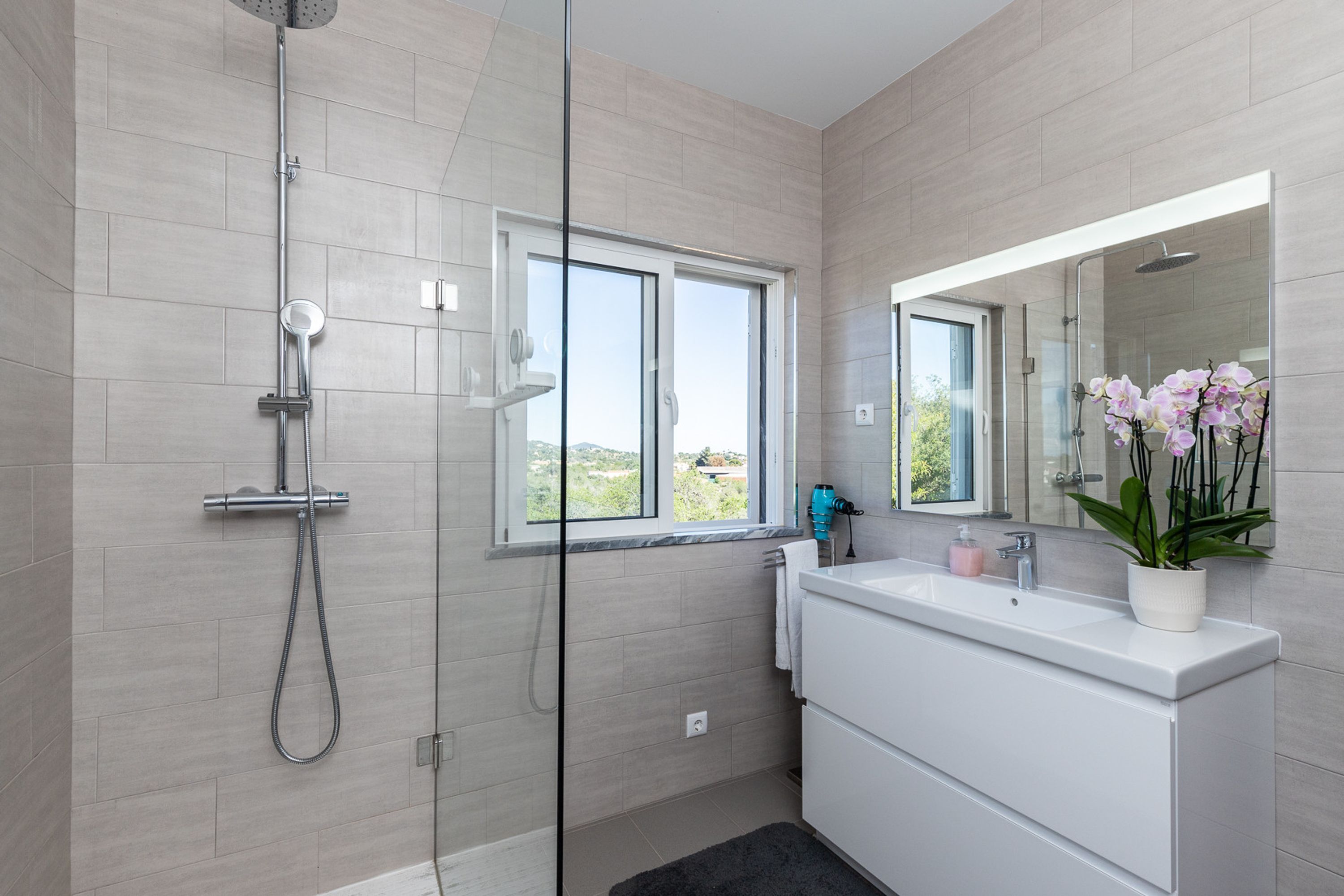 A modern bathroom with a glass-enclosed shower, a large window, a white vanity, and a potted orchid.