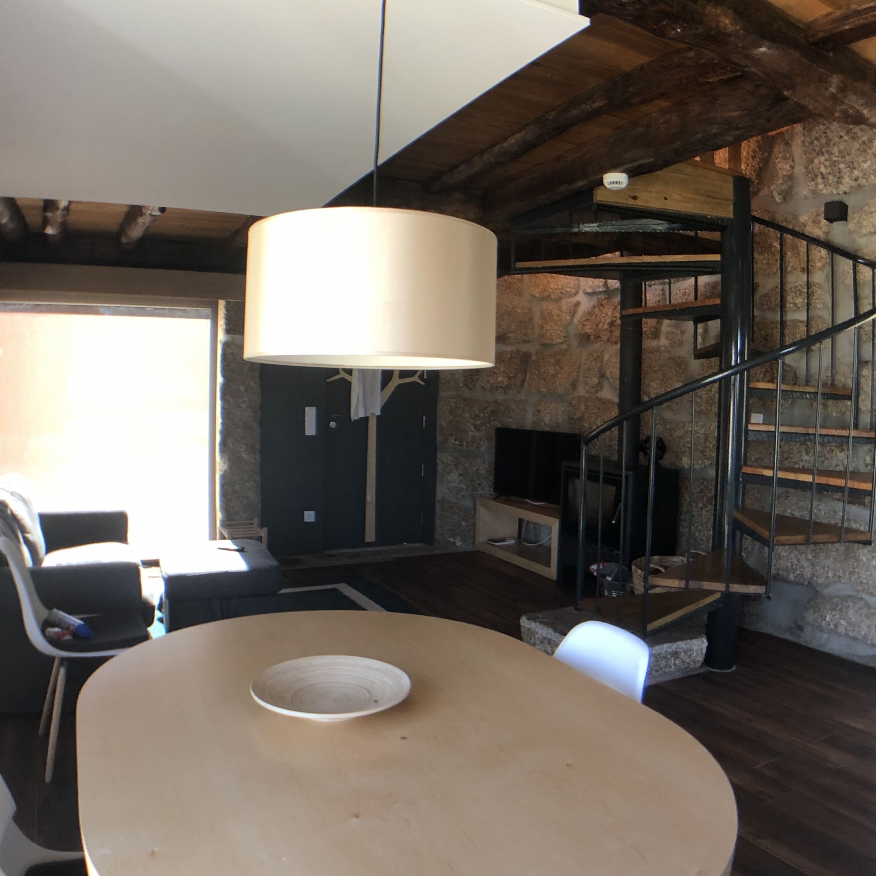 A rustic interior with a round table, a hanging light fixture, a staircase, and a stone wall.