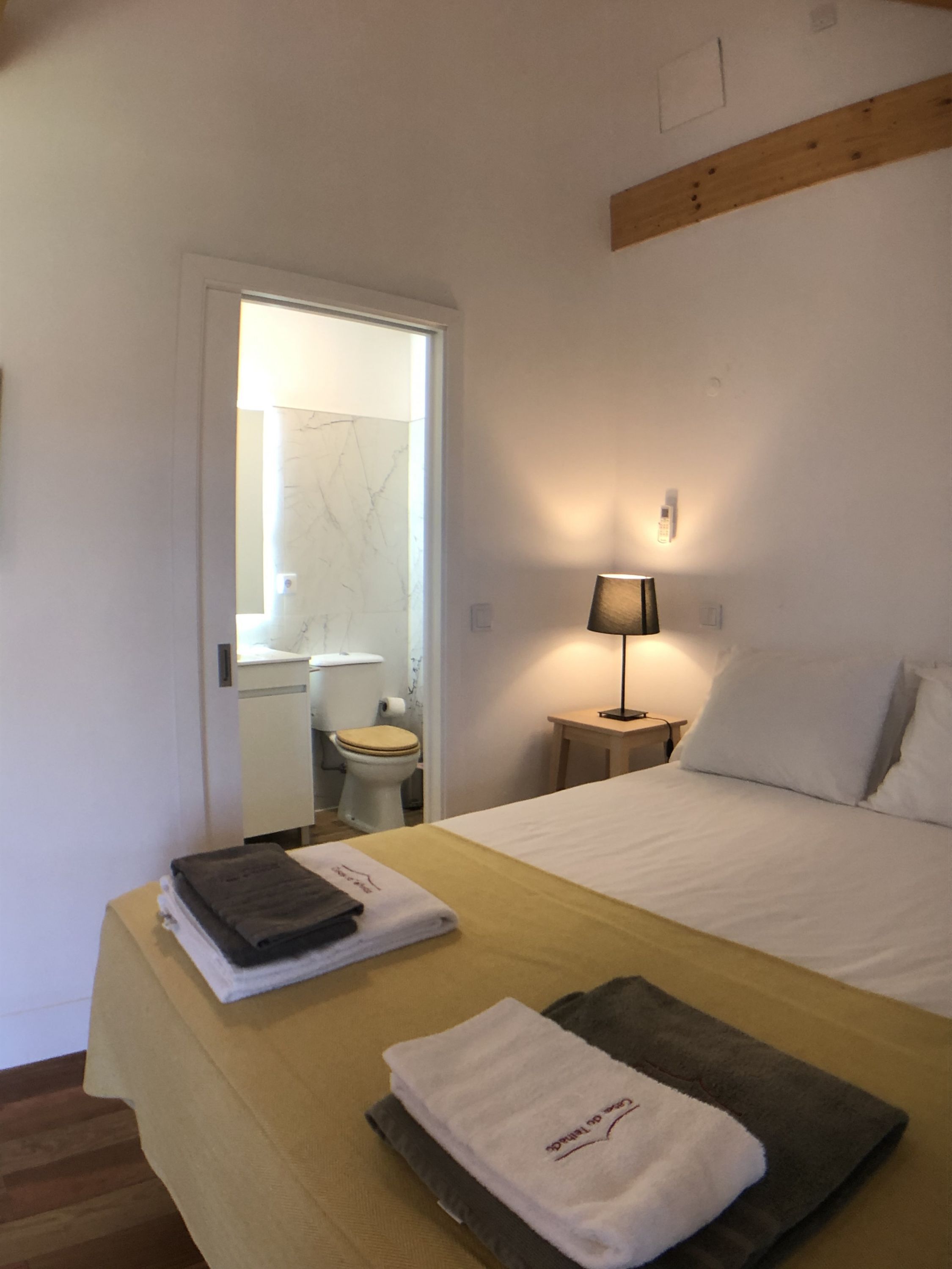 Habitación de hotel con cama, mesita de noche con lámpara, baño visible a través de una puerta abierta y toallas sobre la cama.