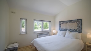 A bedroom with a large bed, two windows, a radiator, a bedside table with a lamp, and a bench. - Vila Beiral in Penafiel (Penafiel)