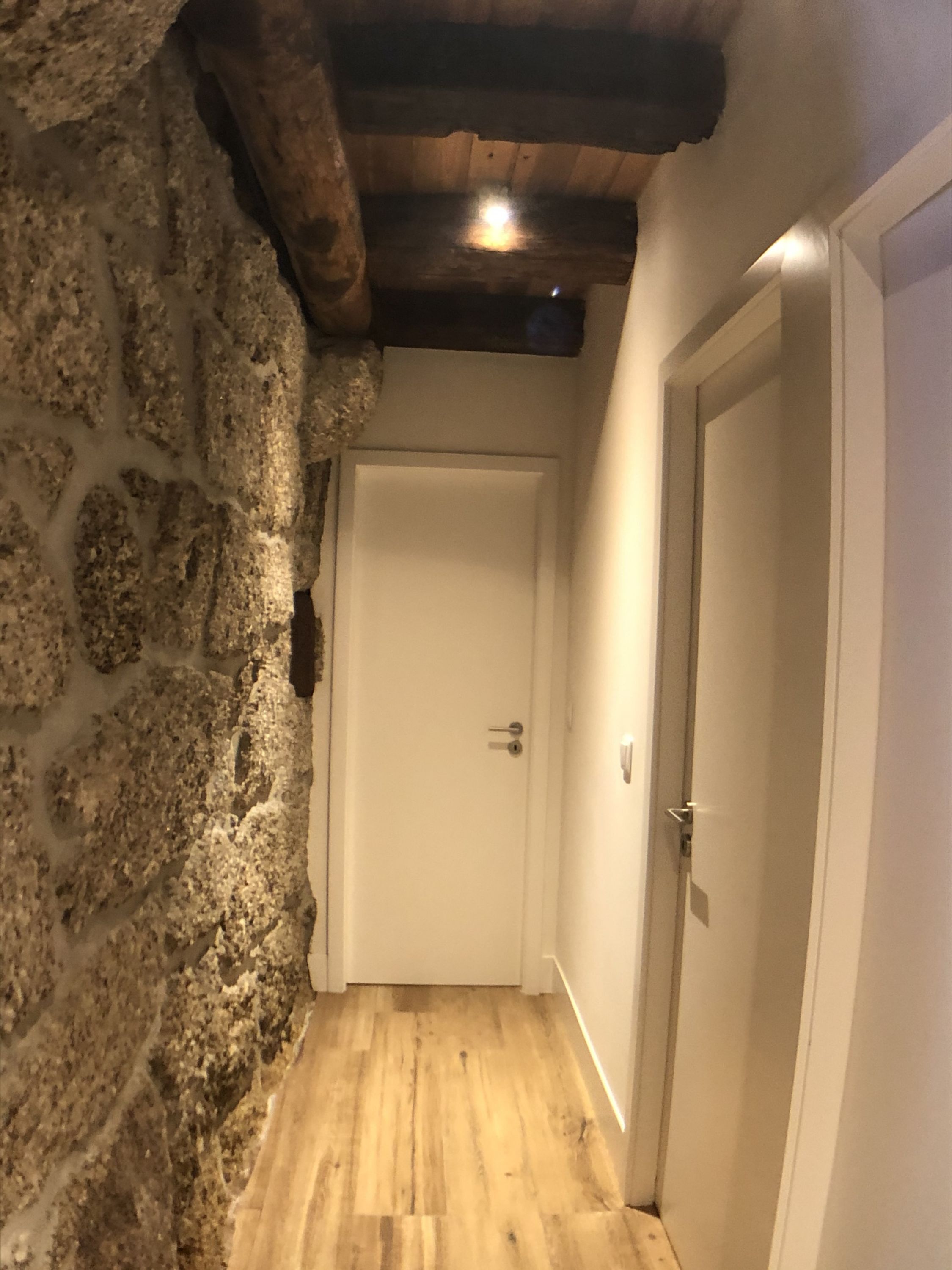 A narrow hallway with a stone wall on one side and a wooden floor. There is a white door at the end of the hallway.