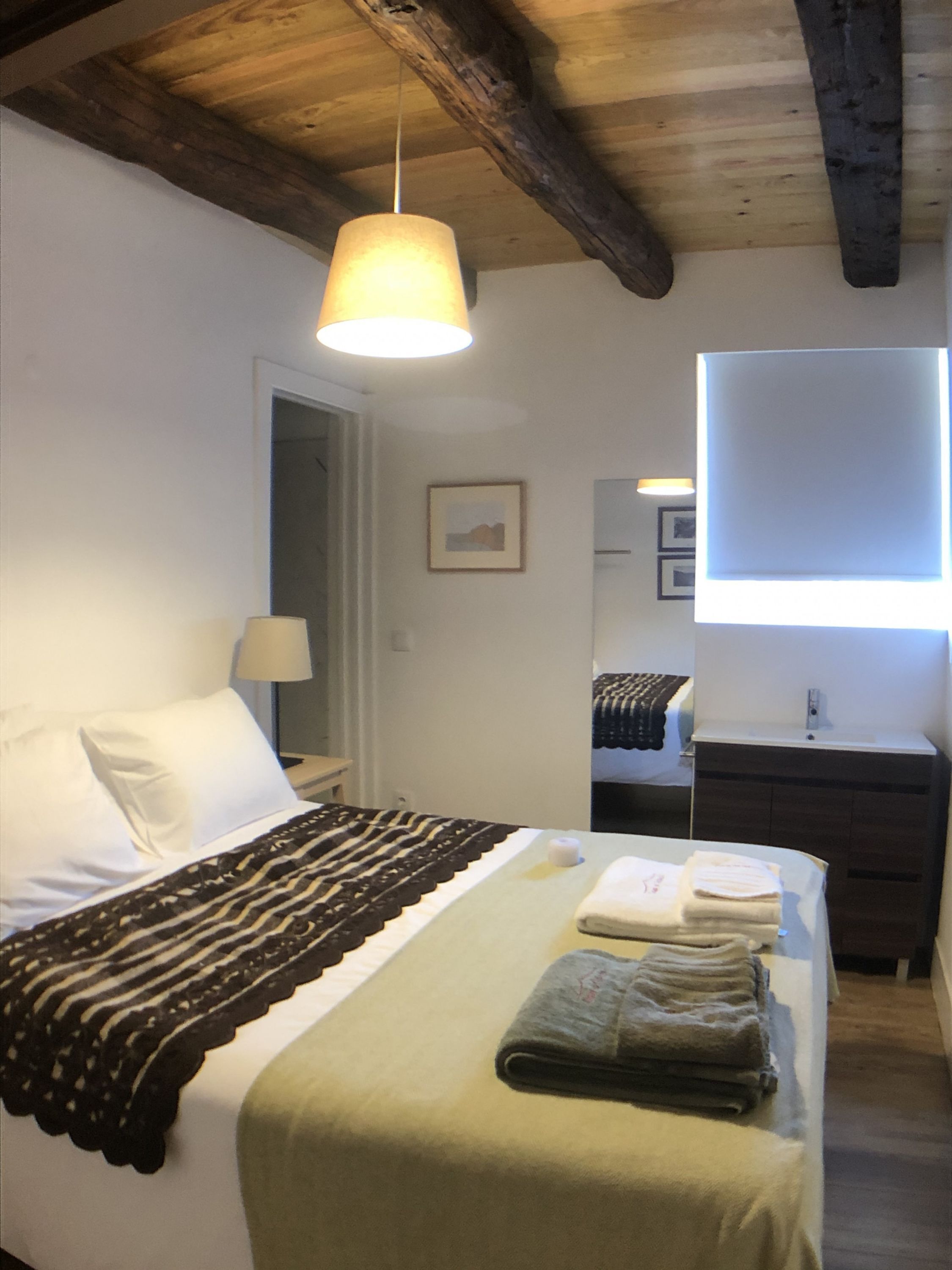 Une chambre à coucher comprenant un lit, une lampe de chevet, un luminaire suspendu, une commode et une fenêtre avec des stores.