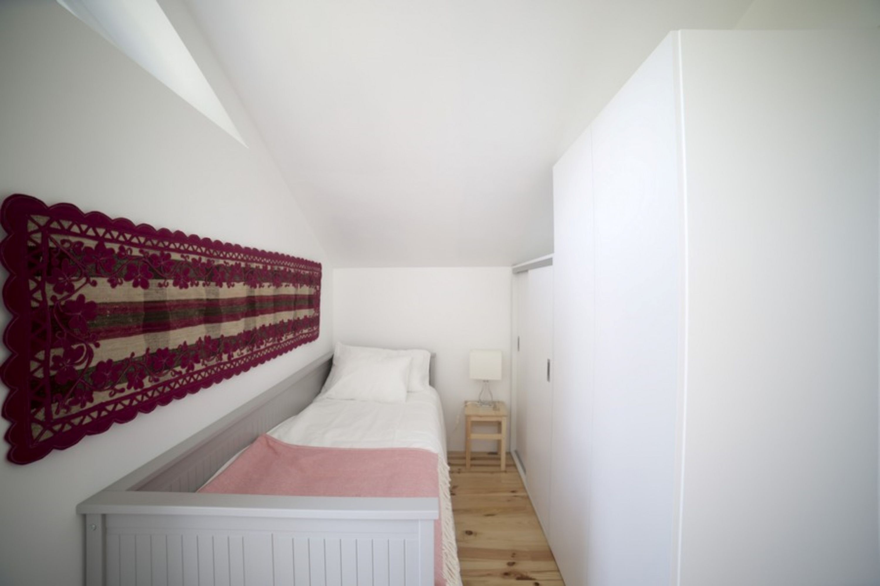 A narrow, white-walled room with a single bed, a small wooden nightstand, and a decorative wall hanging.