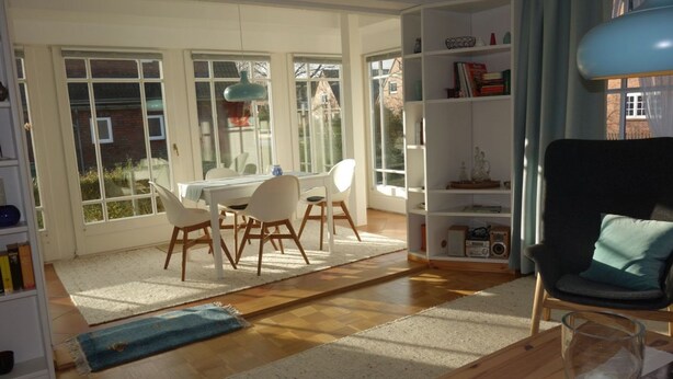 A bright living room with a dining area, large windows, and a bookshelf.