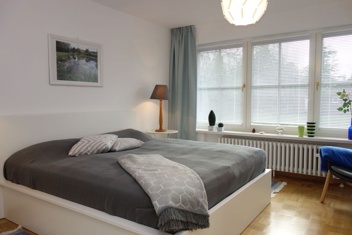 Ett sovrum med en stor säng, en sänglampa, ett fönster med persienner, ett element och ett litet bord med en växt.