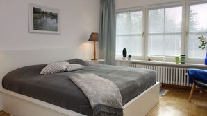 A bedroom with a large bed, a bedside lamp, a window with blinds, a radiator, and a small table with a plant.