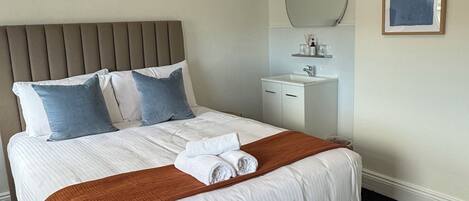 A neatly made bed with white and blue pillows, a brown throw blanket, and rolled towels.