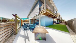 A patio area with a wooden table and chairs, a covered seating area, and a green lawn. - OV34 Shared Pool, Close to the Beach, Ping Pong Table, Firepit, Putting Green (Port Aransas)