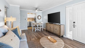 Una sala moderna con sofá, mesa de centro y televisor. Se ve un comedor con sillas y una cocina al fondo.