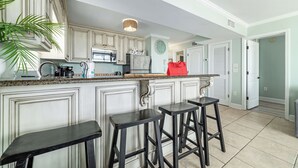 A kitchen with white cabinetry, a granite countertop, and three black stools. - Seaside Beach & Racquet Club 3301 – Beachfront w Pool! 🌊☀️ (Orange Beach)