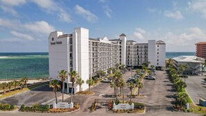 A multi-story hotel with a white facade, named Grand Pointe, featuring a parking lot with several cars, palm trees, and a view of the ocean. - BEACHFRONT, FAMILY FRIENDLY, LOW FLOOR, INDOOR/OUTDOOR POOL | BEACHBALL PROPERTIES (Orange Beach)