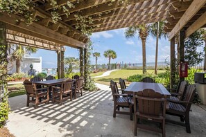 An outdoor patio with wooden tables and chairs, surrounded by greenery and palm trees. - BEACHFRONT, FAMILY FRIENDLY, LOW FLOOR, INDOOR/OUTDOOR POOL | BEACHBALL PROPERTIES (Orange Beach)